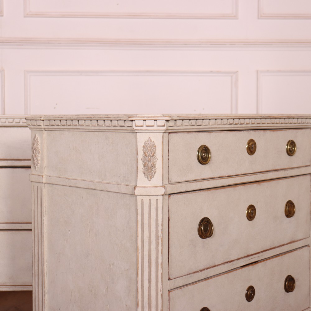 A pair of Swedish Gustavian style off-white painted commodes featuring three drawers each, brass hardware, fluted sides, and square tapered feet. View VI