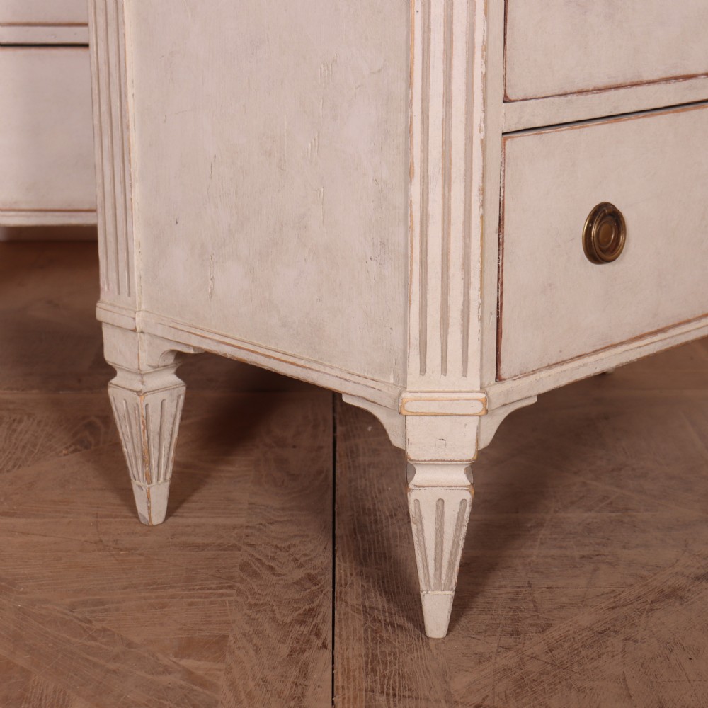 A pair of Swedish Gustavian style off-white painted commodes featuring three drawers each, brass hardware, fluted sides, and square tapered feet. View VIII