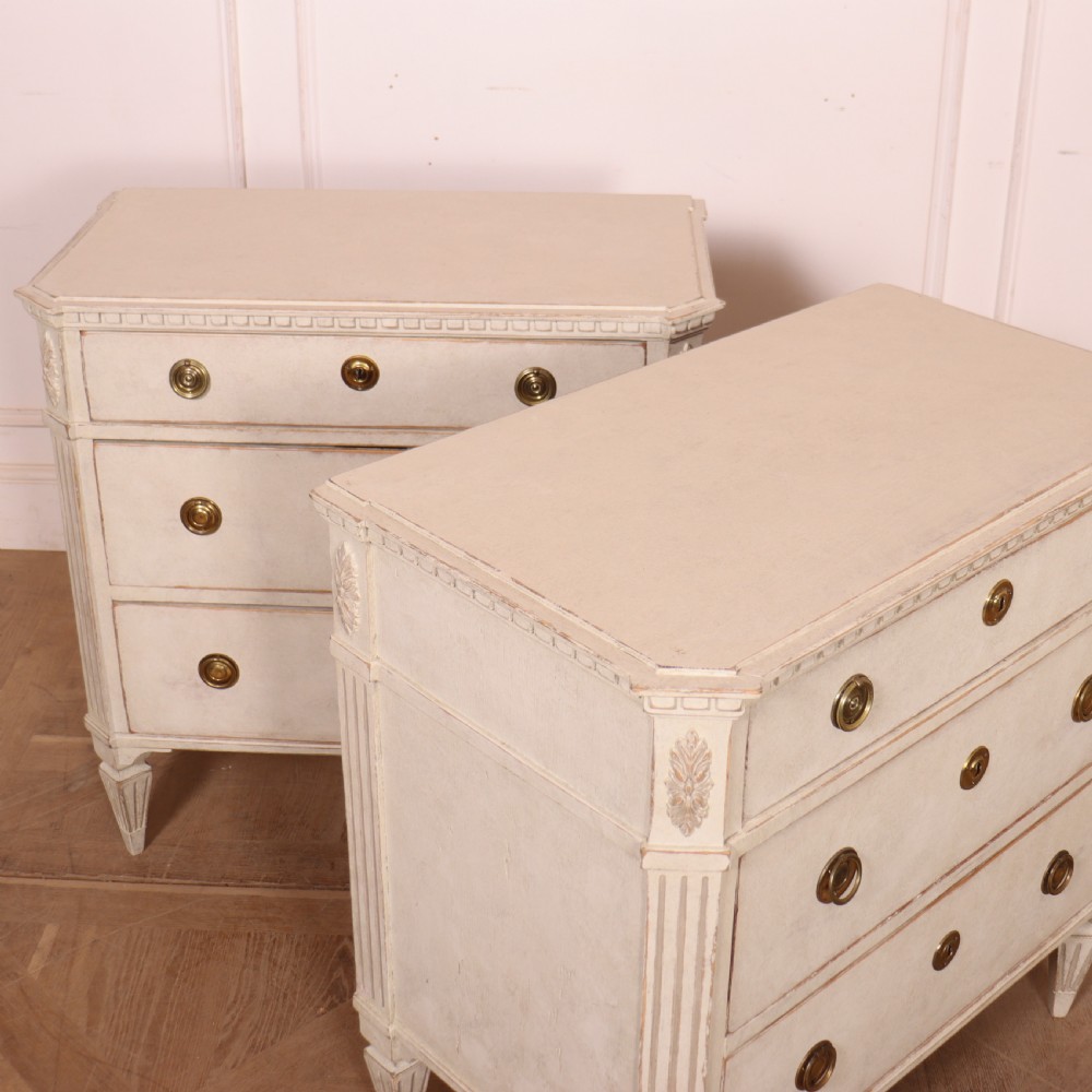 A pair of Swedish Gustavian style off-white painted commodes featuring three drawers each, brass hardware, fluted sides, and square tapered feet. View IX