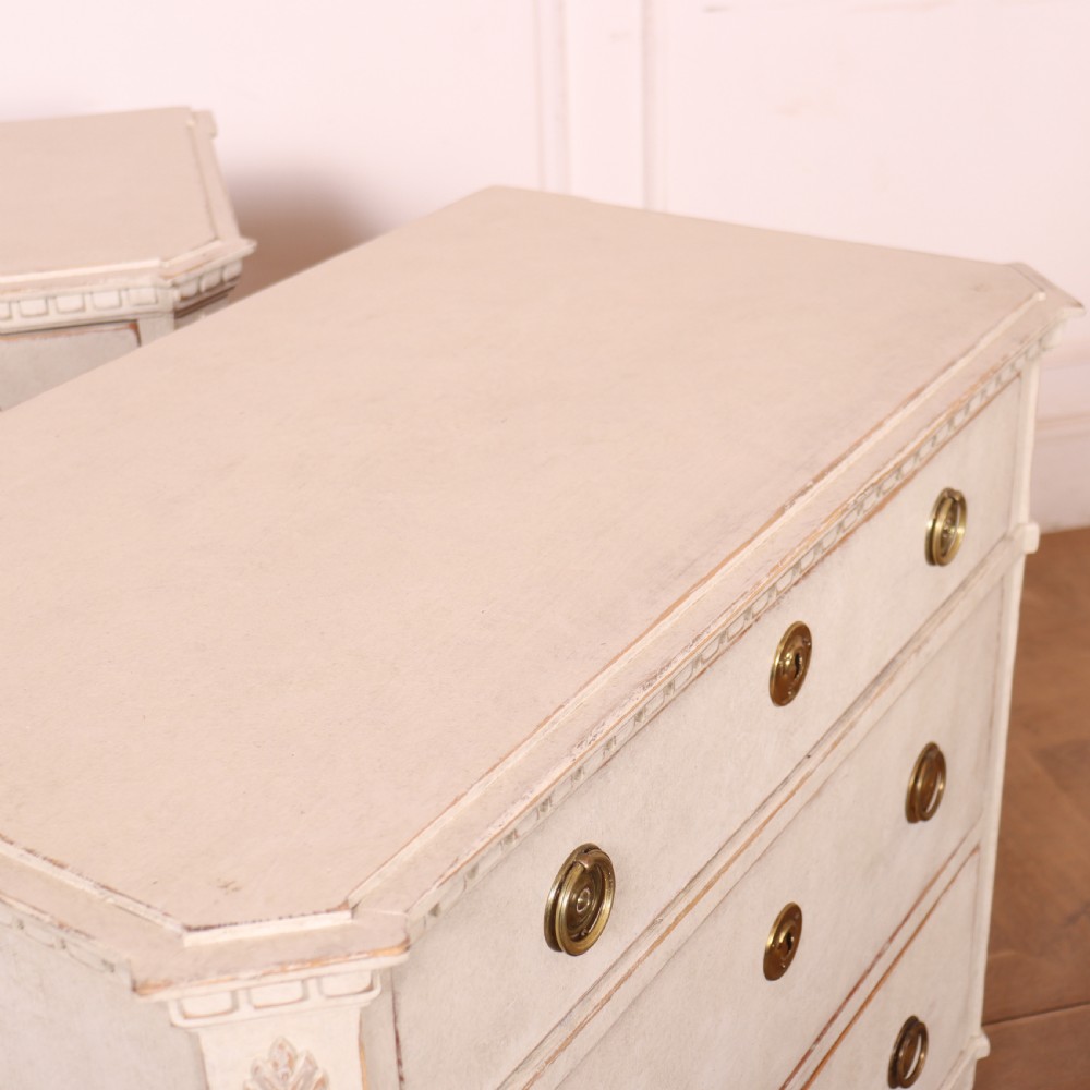 A pair of Swedish Gustavian style off-white painted commodes featuring three drawers each, brass hardware, fluted sides, and square tapered feet. View X