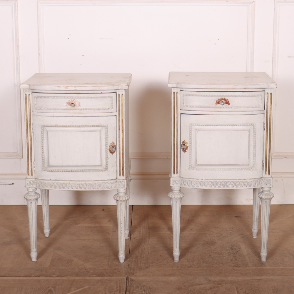 A pair of off-white painted Swedish Gustavian style bedside cupboards featuring fluted tapered legs, gilded trim, top drawers, and cupboard doors.