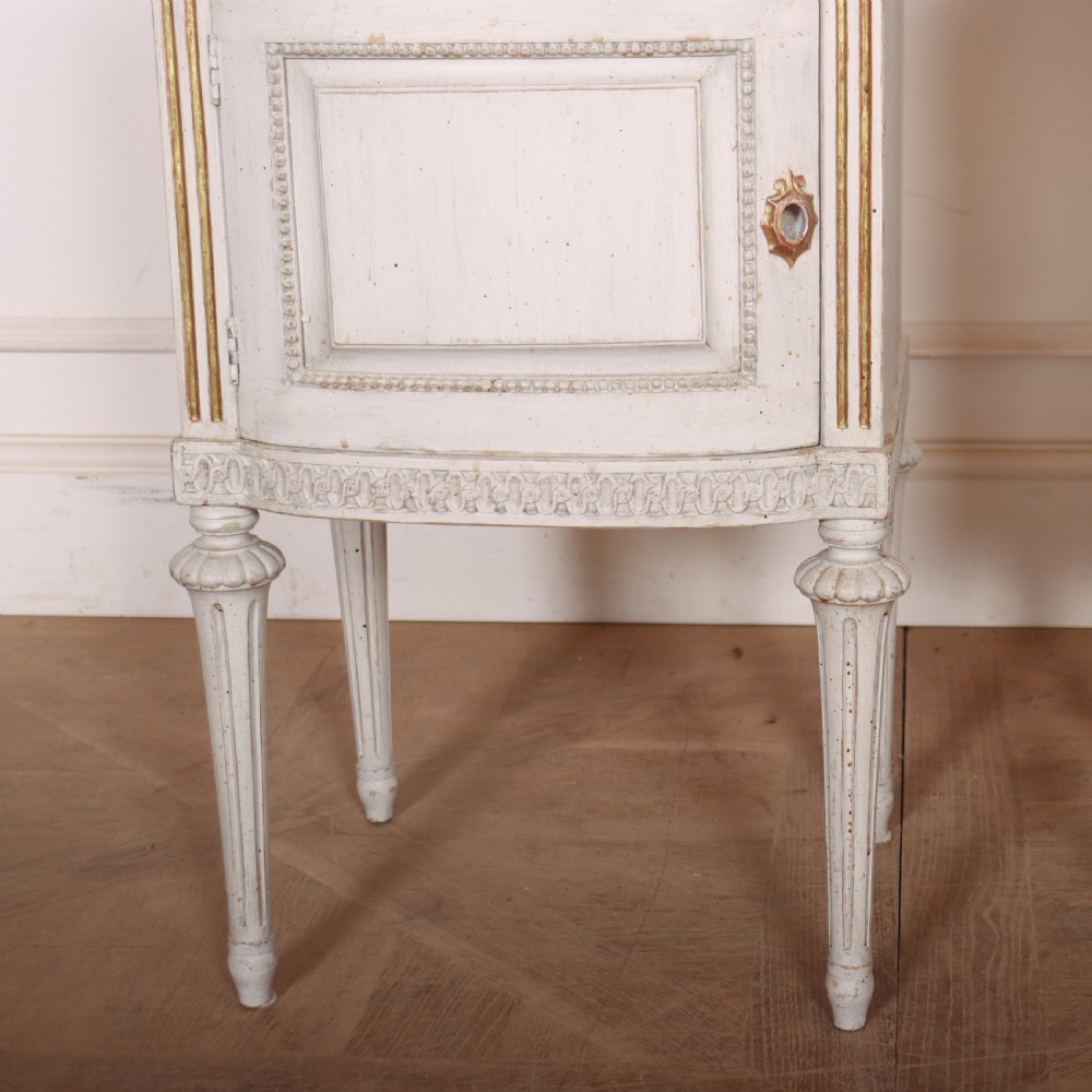 A pair of off-white painted Swedish Gustavian style bedside cupboards featuring fluted tapered legs, gilded trim, top drawers, and cupboard doors. View III