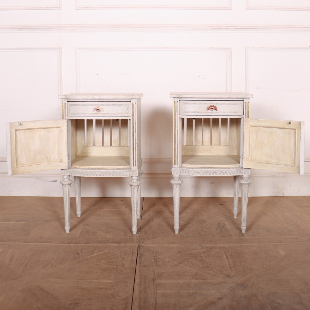 A pair of off-white painted Swedish Gustavian style bedside cupboards featuring fluted tapered legs, gilded trim, top drawers, and cupboard doors. View V