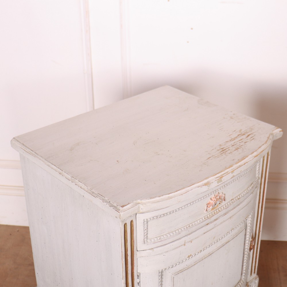 A pair of off-white painted Swedish Gustavian style bedside cupboards featuring fluted tapered legs, gilded trim, top drawers, and cupboard doors. View VII