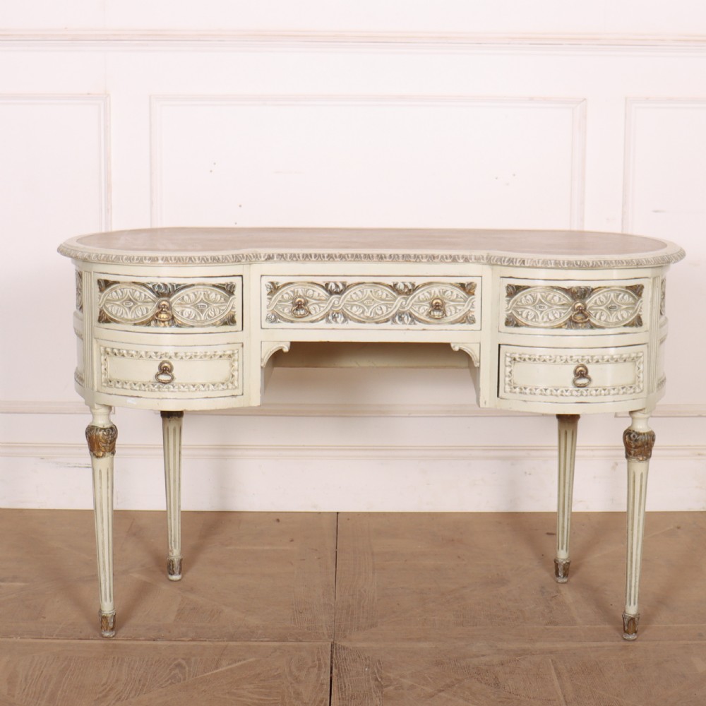 An antique cream-painted curved writing desk featuring carved neoclassical scrollwork, brass drop-ring handles, and tapered fluted legs.