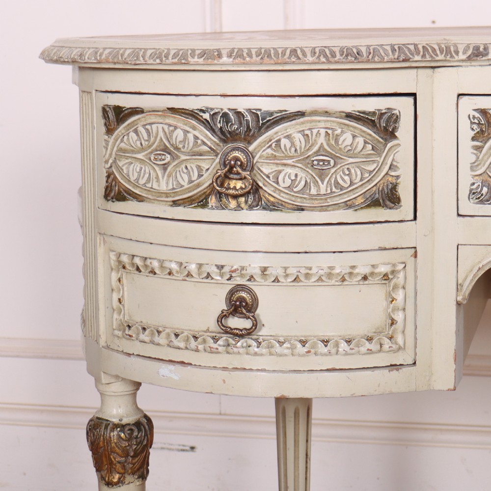 An antique cream-painted curved writing desk featuring carved neoclassical scrollwork, brass drop-ring handles, and tapered fluted legs. View II