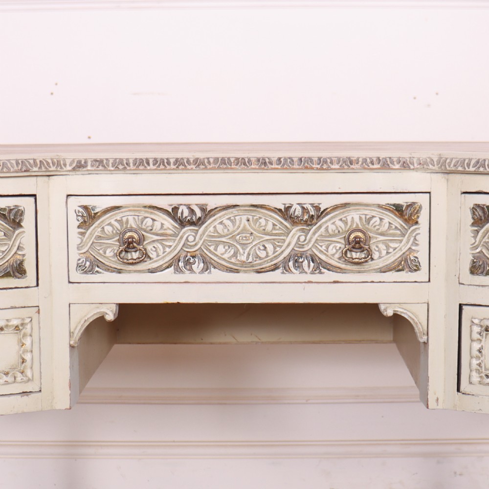 An antique cream-painted curved writing desk featuring carved neoclassical scrollwork, brass drop-ring handles, and tapered fluted legs. View III