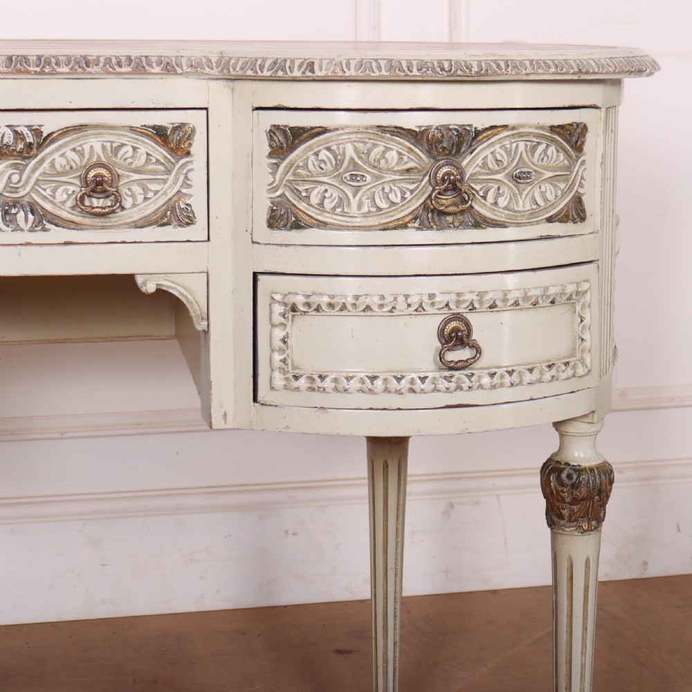 An antique cream-painted curved writing desk featuring carved neoclassical scrollwork, brass drop-ring handles, and tapered fluted legs. View IV