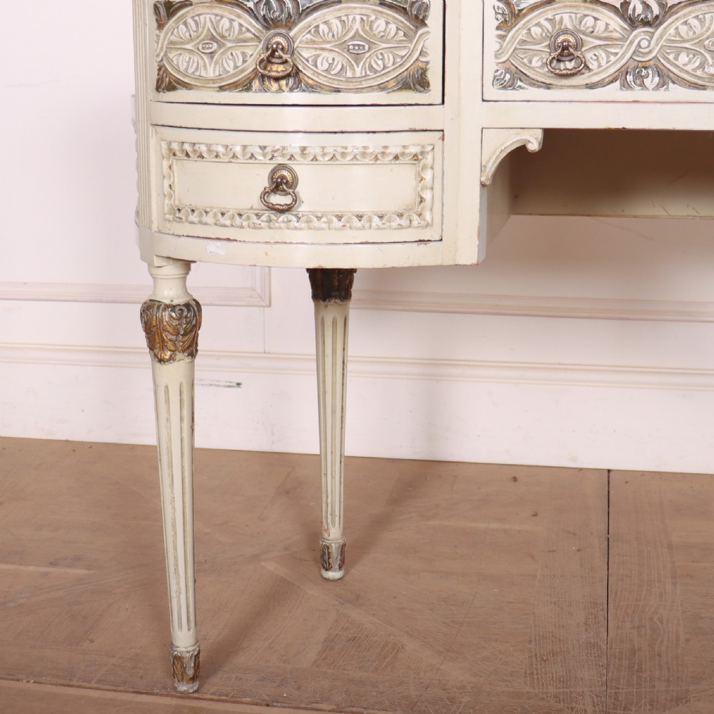 An antique cream-painted curved writing desk featuring carved neoclassical scrollwork, brass drop-ring handles, and tapered fluted legs. View VII