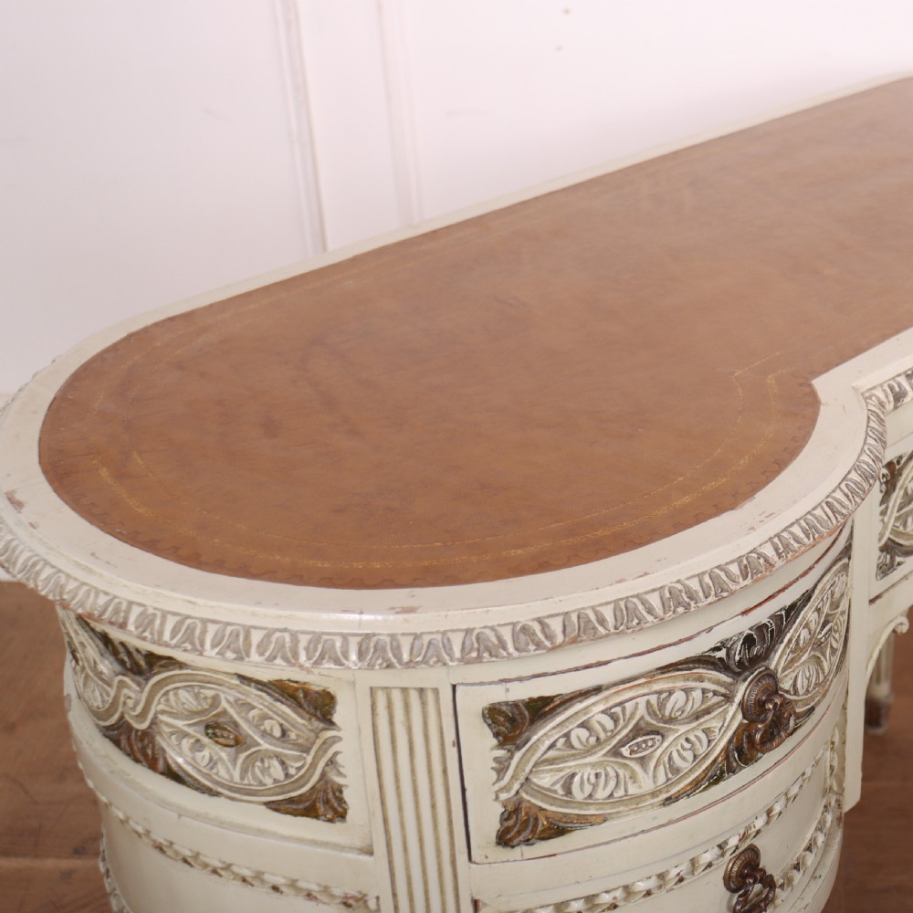 An antique cream-painted curved writing desk featuring carved neoclassical scrollwork, brass drop-ring handles, and tapered fluted legs. View IX