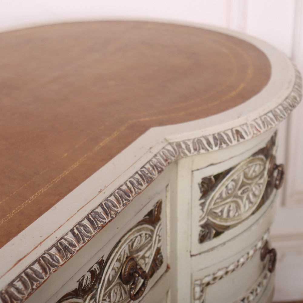 An antique cream-painted curved writing desk featuring carved neoclassical scrollwork, brass drop-ring handles, and tapered fluted legs. View X