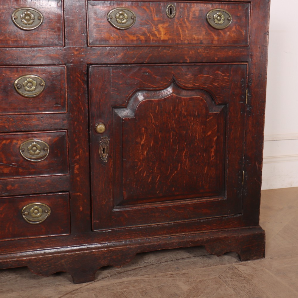 An eighteenth-century dark oak dresser base featuring three top drawers and a central column of drawers flanked by two arched fielded panel cupboard doors. View V