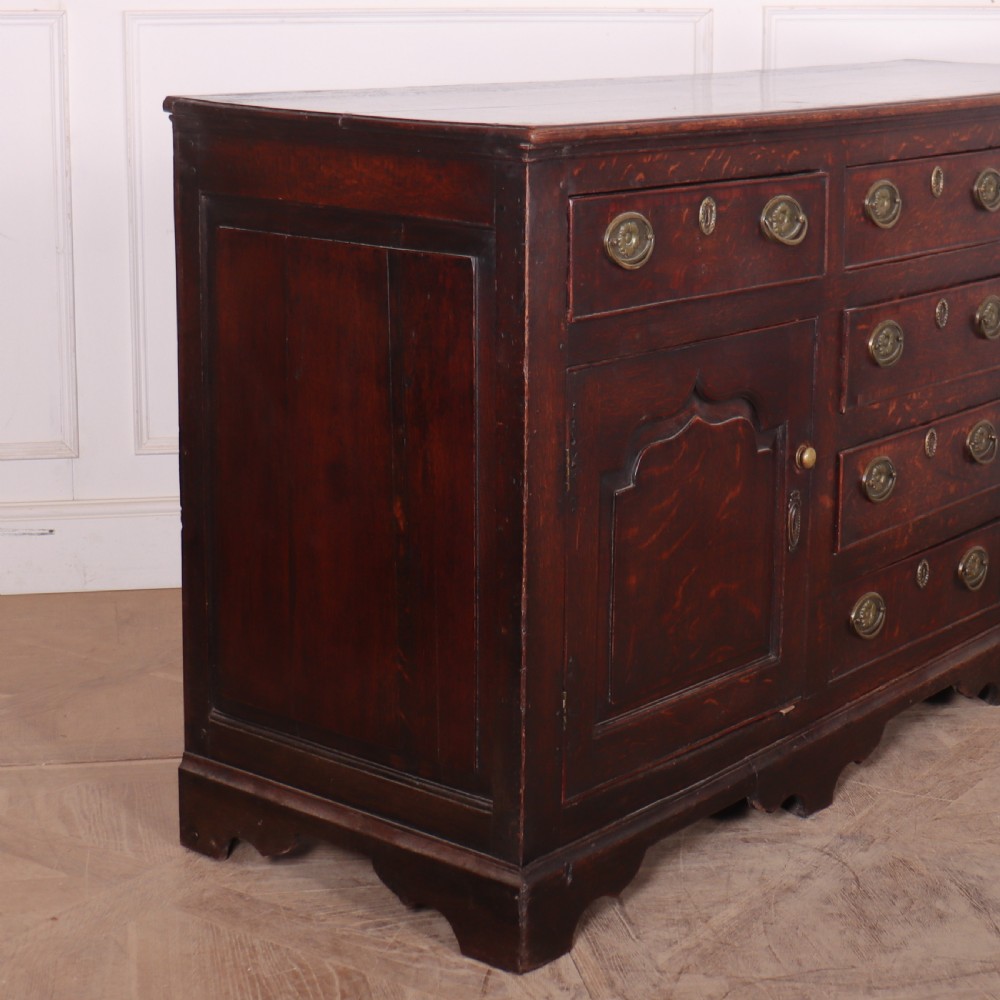 An eighteenth-century dark oak dresser base featuring three top drawers and a central column of drawers flanked by two arched fielded panel cupboard doors. View IX