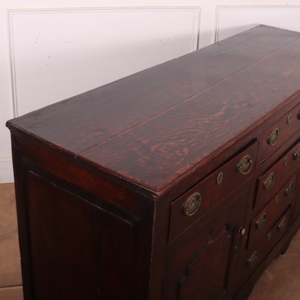 An eighteenth-century dark oak dresser base featuring three top drawers and a central column of drawers flanked by two arched fielded panel cupboard doors. View X