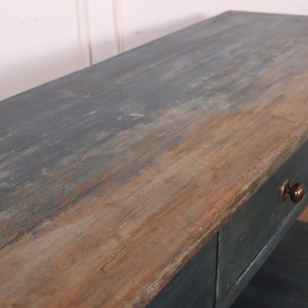 A long muted blue painted wooden dresser base featuring four drawers with brass knobs, a exposed wood top surface, and an open slatted lower shelf. View X