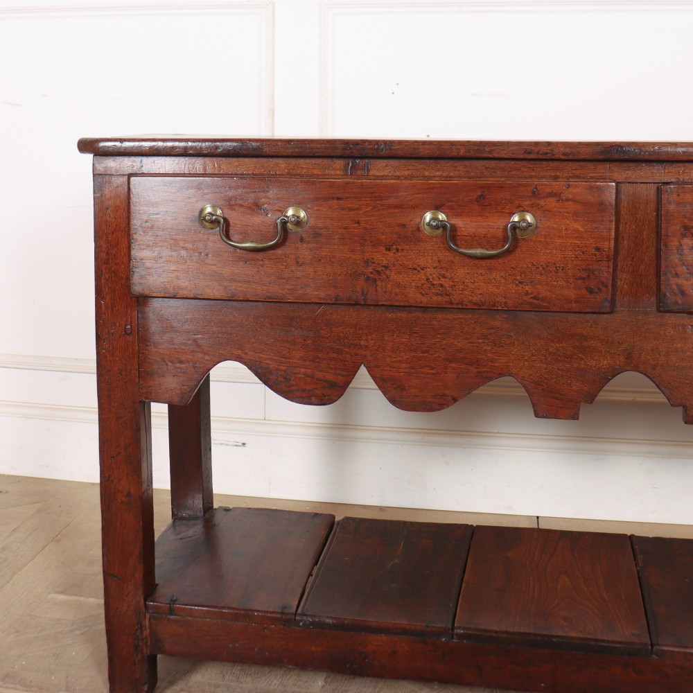 An antique dark oak Welsh dresser base featuring two drawers with brass swan-neck handles, a scalloped apron, and a lower potboard shelf. View II