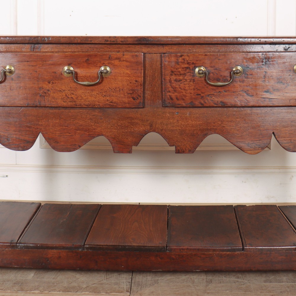 An antique dark oak Welsh dresser base featuring two drawers with brass swan-neck handles, a scalloped apron, and a lower potboard shelf. View IV