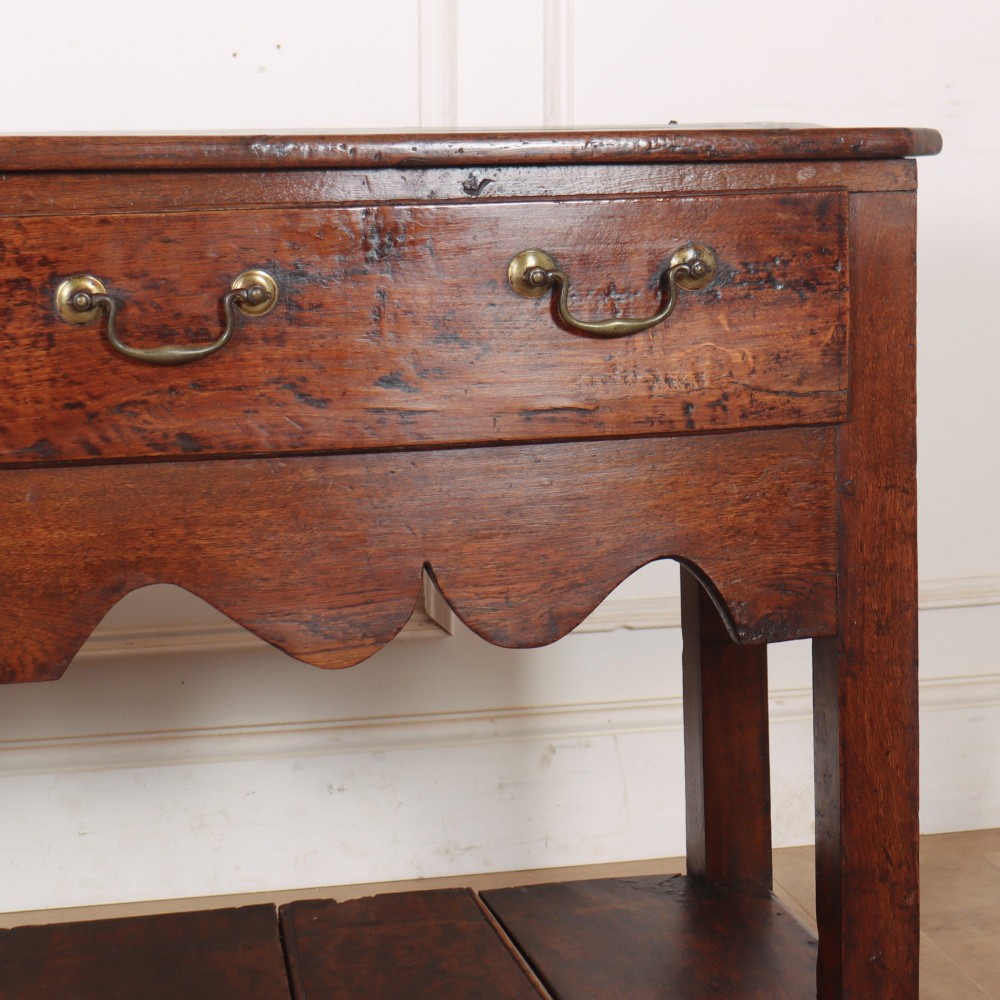 An antique dark oak Welsh dresser base featuring two drawers with brass swan-neck handles, a scalloped apron, and a lower potboard shelf. View V
