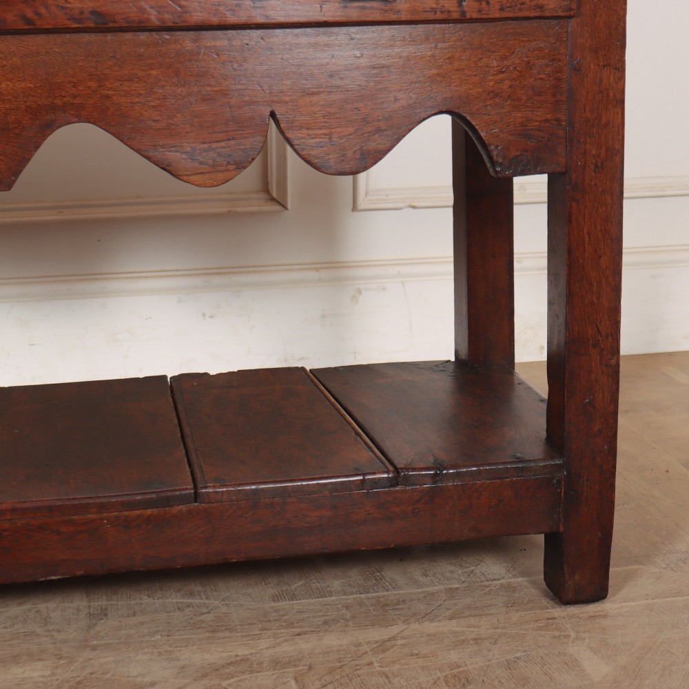 An antique dark oak Welsh dresser base featuring two drawers with brass swan-neck handles, a scalloped apron, and a lower potboard shelf. View VI