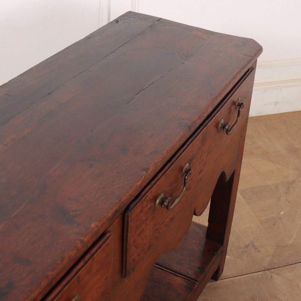 An antique dark oak Welsh dresser base featuring two drawers with brass swan-neck handles, a scalloped apron, and a lower potboard shelf. View IX