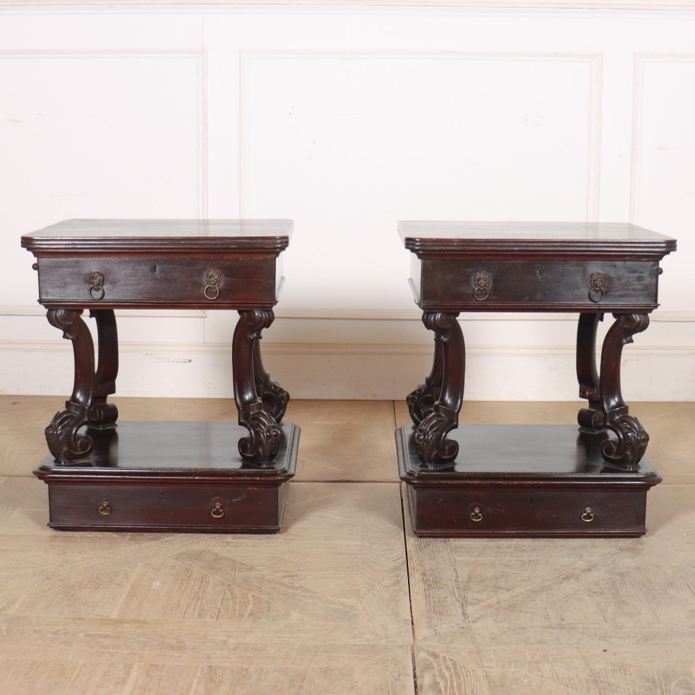 A pair of dark mahogany bedside tables featuring carved scroll supports, upper and lower drawers with brass drop-ring handles, resting on solid plinth bases.