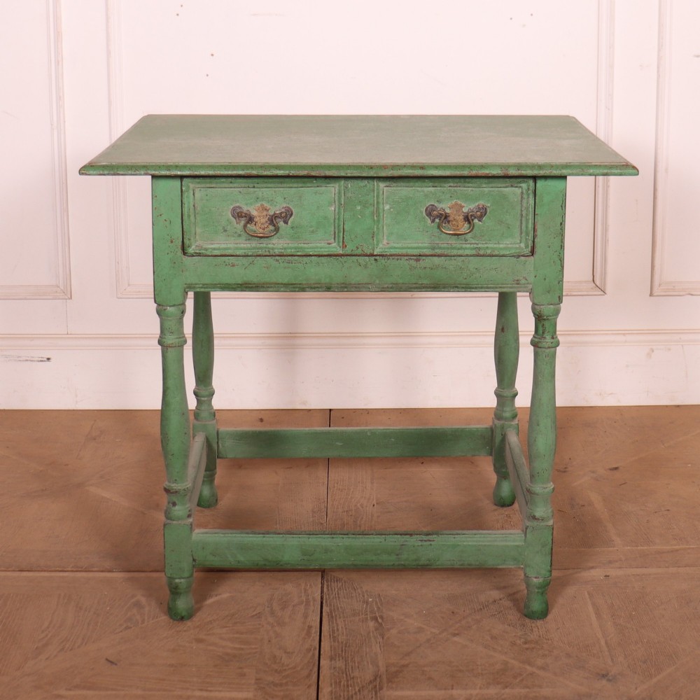 An antique English oak lamp table featuring a distressed green painted finish, two drawers with brass handles, turned legs, and a lower stretcher base.