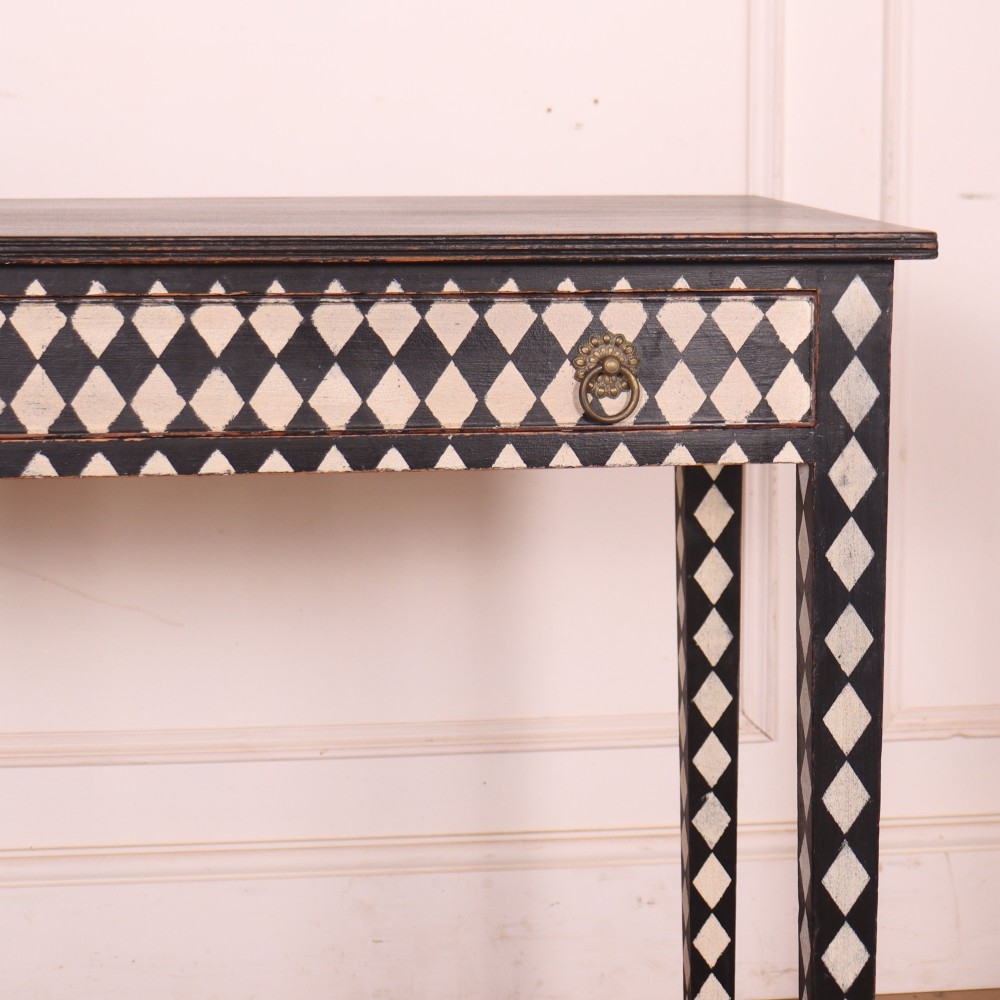 A rectangular black painted lamp table featuring a hand-painted cream and black harlequin diamond pattern on the front drawer and square legs, finished with two brass ring pull handles. View III