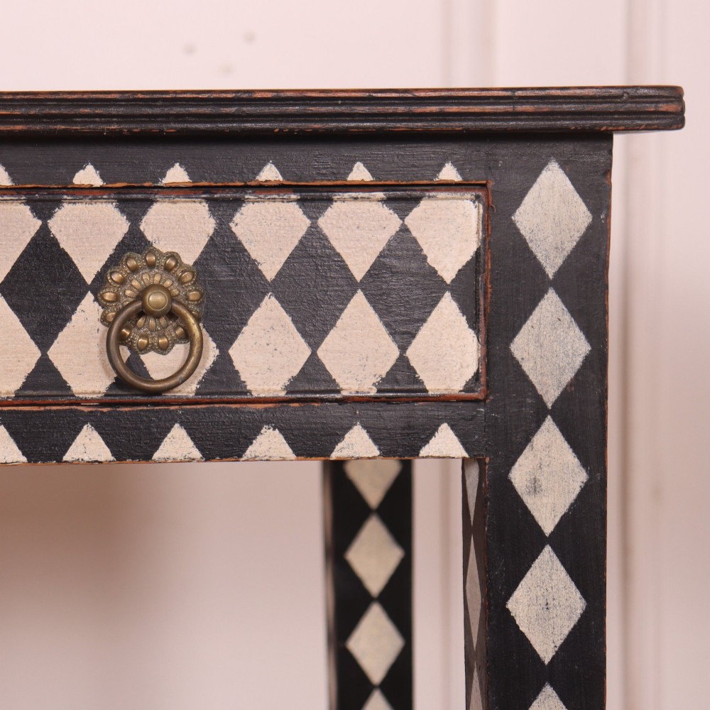 A rectangular black painted lamp table featuring a hand-painted cream and black harlequin diamond pattern on the front drawer and square legs, finished with two brass ring pull handles. View VI