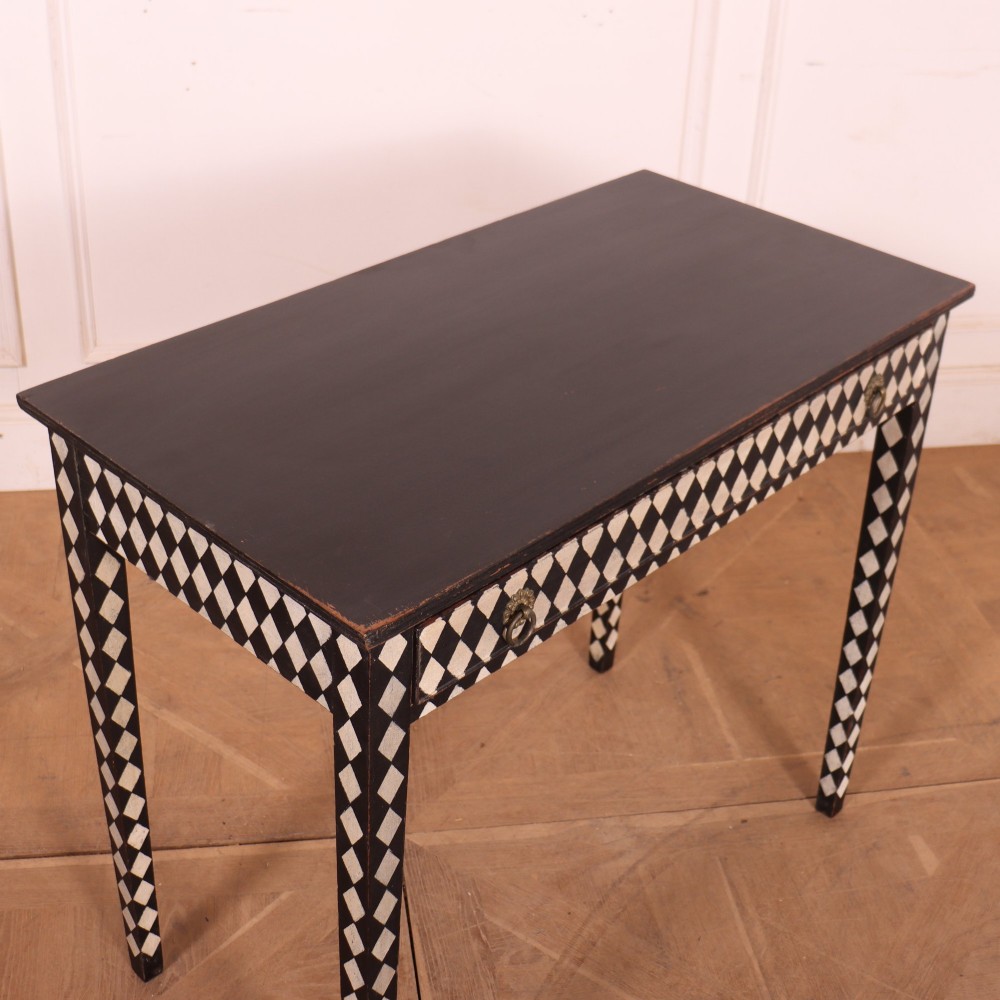 A rectangular black painted lamp table featuring a hand-painted cream and black harlequin diamond pattern on the front drawer and square legs, finished with two brass ring pull handles. View VII