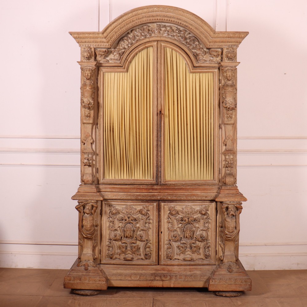 A large Italian carved wood vitrine featuring arched upper glazed doors lined with fabric, flanked by caryatid figures and intricate relief panels.