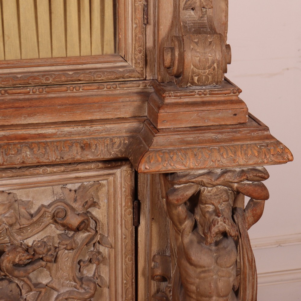 A large Italian carved wood vitrine featuring arched upper glazed doors lined with fabric, flanked by caryatid figures and intricate relief panels. View VI