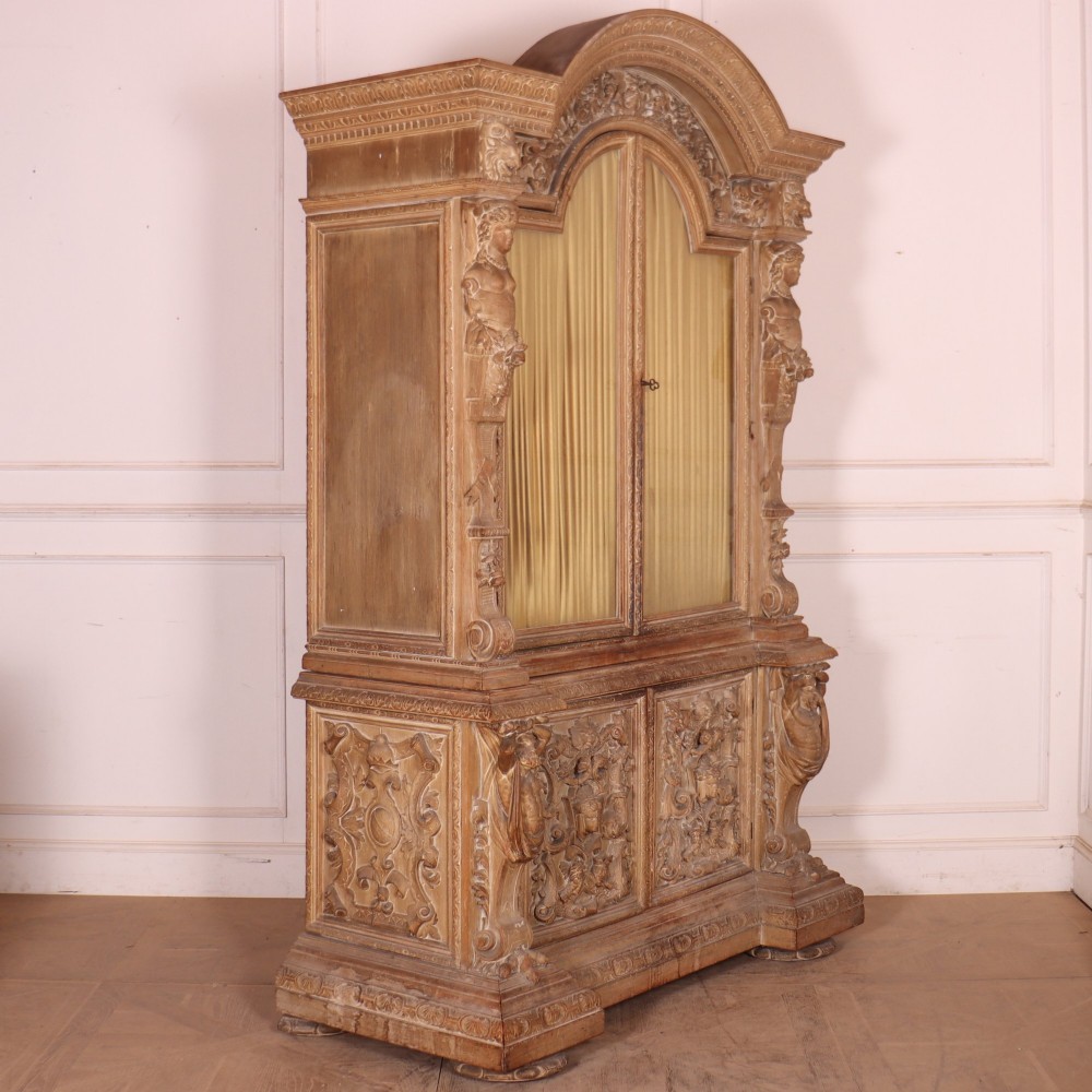 A large Italian carved wood vitrine featuring arched upper glazed doors lined with fabric, flanked by caryatid figures and intricate relief panels. View XII