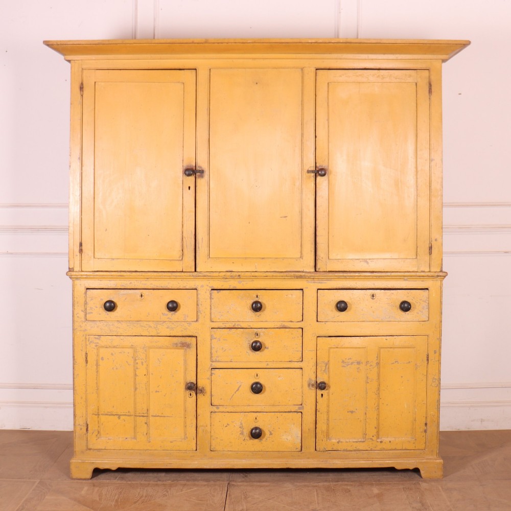 A large antique yellow-painted housekeeper cupboard featuring three upper cupboard doors above an arrangement of drawers and lower cupboards.