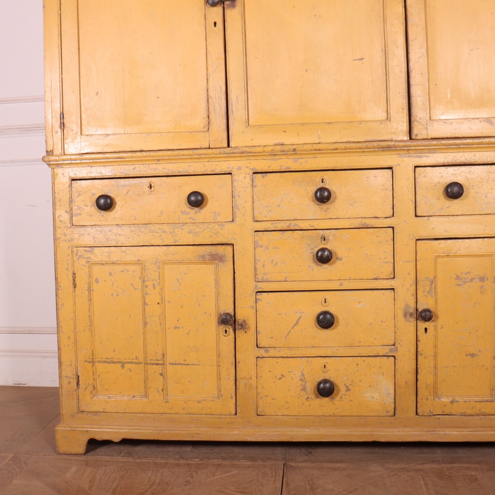 A large antique yellow-painted housekeeper cupboard featuring three upper cupboard doors above an arrangement of drawers and lower cupboards. View V