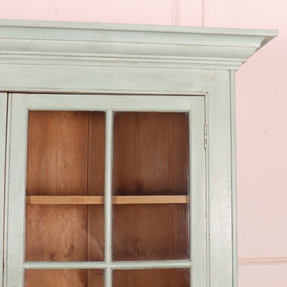 An English country painted pine dresser featuring two glazed upper doors revealing natural wood shelves above two panelled lower doors in a pale green finish. View III