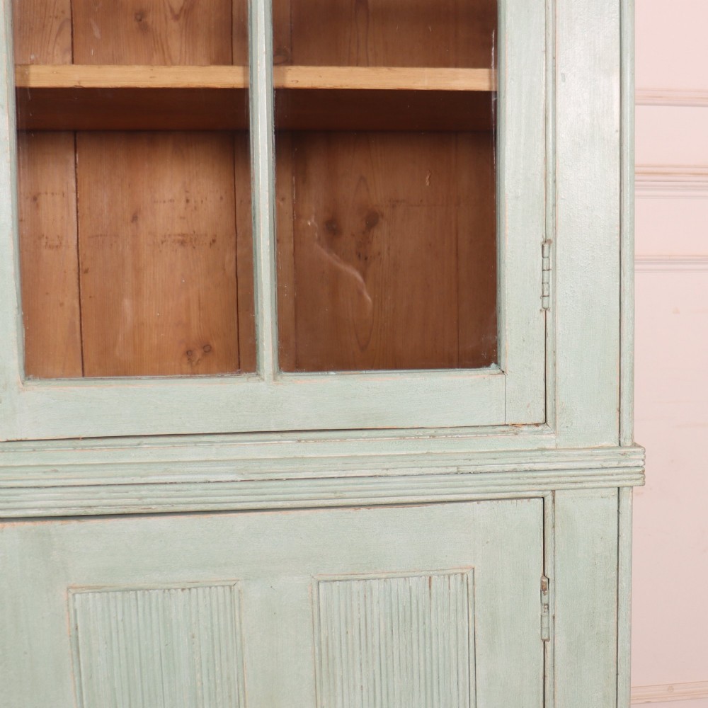 An English country painted pine dresser featuring two glazed upper doors revealing natural wood shelves above two panelled lower doors in a pale green finish. View IV