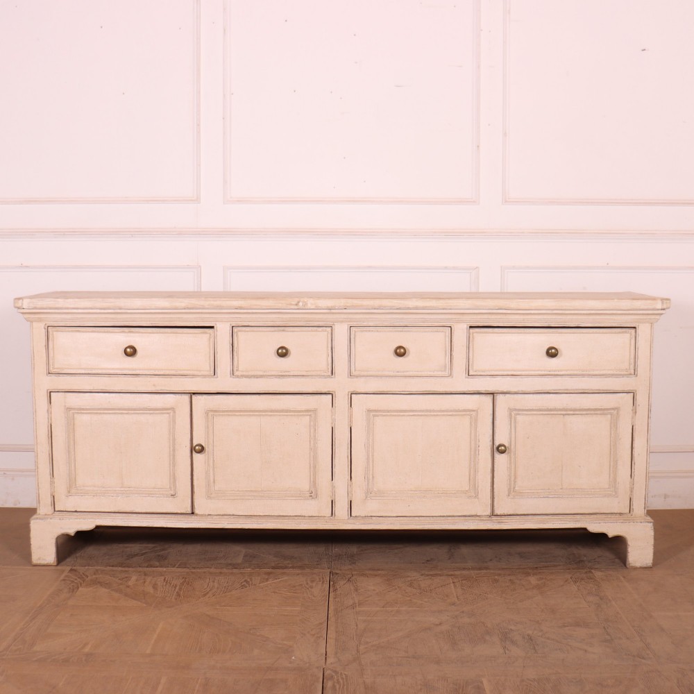 A large off-white painted English dresser base featuring four drawers above four cupboard doors with small brass knob handles.