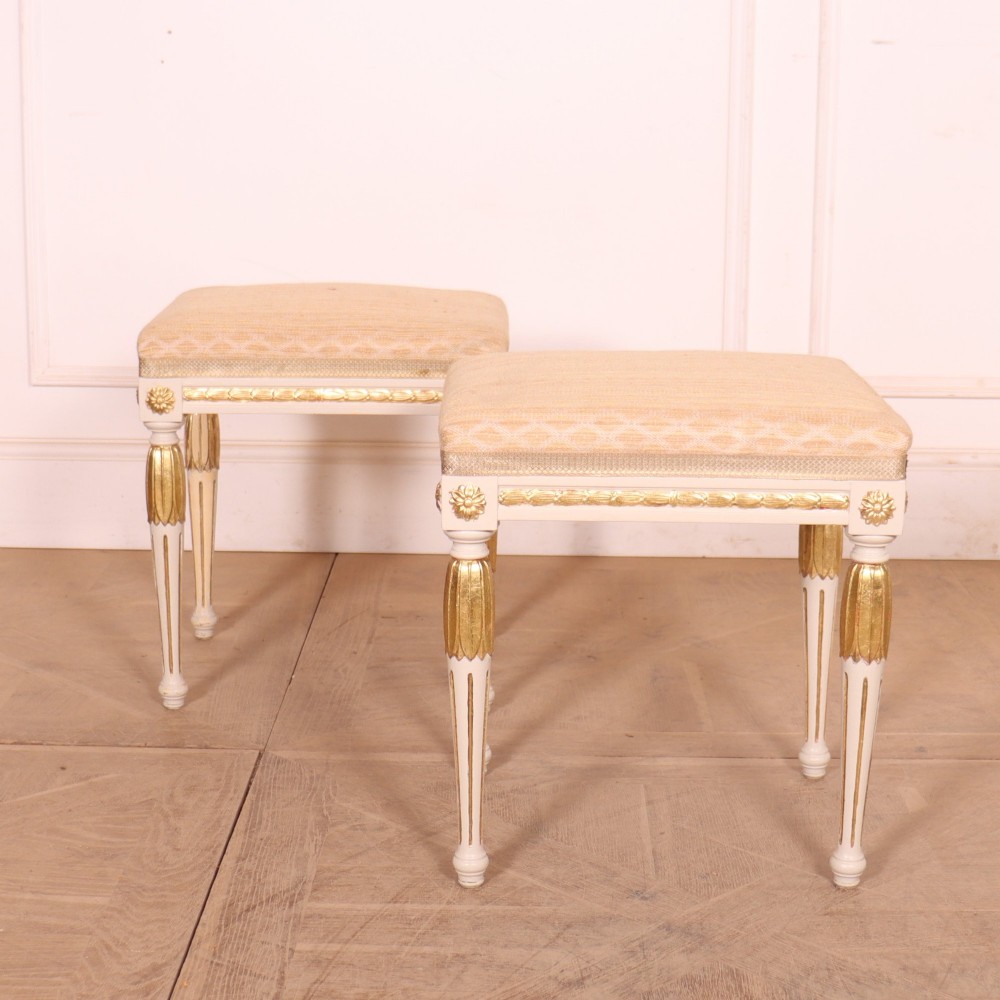A pair of Neoclassical Swedish Gustavian style footstools with white painted legs, gold leaf fluted details, and cream patterned upholstered seats.