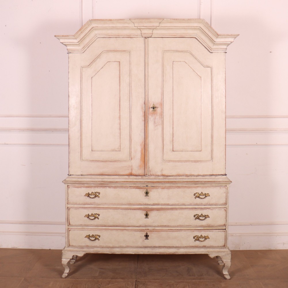 A tall antique Swedish painted linen cupboard featuring shaped panel doors over three drawers with brass handles in an off-white distressed finish.