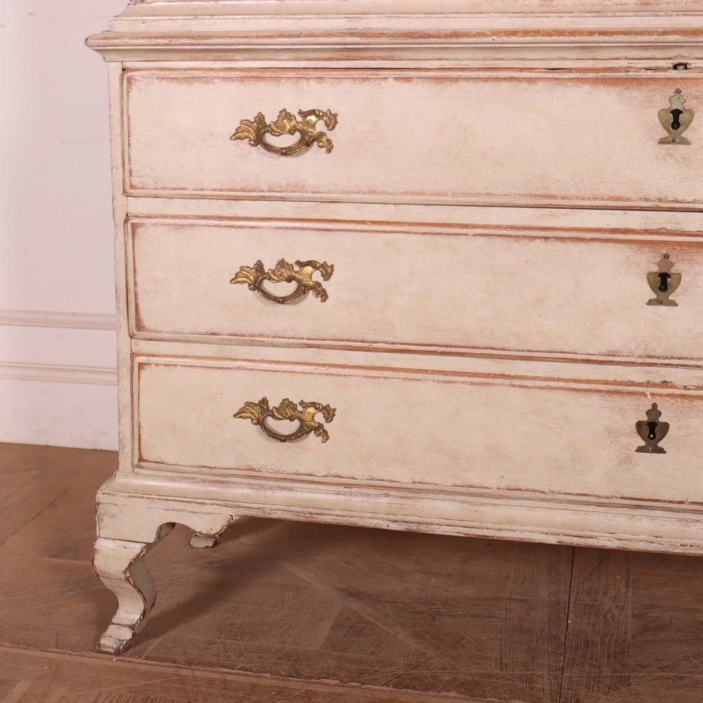 A tall antique Swedish painted linen cupboard featuring shaped panel doors over three drawers with brass handles in an off-white distressed finish. View V