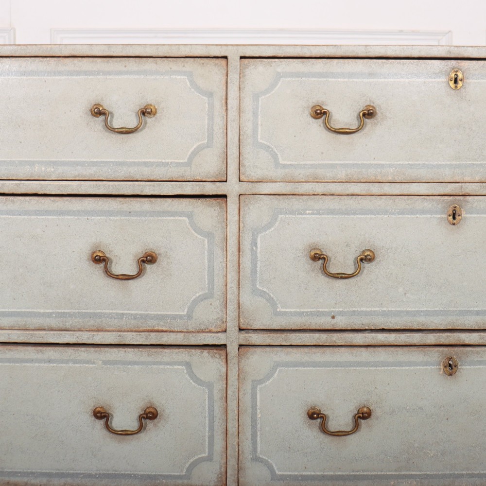 A wide pale grey painted chest featuring six drawers decorated with blue painted border outlines and brass drop handles. View IV