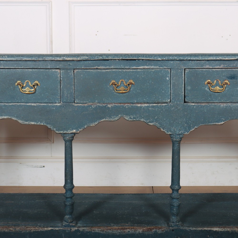 A long blue-painted wooden dresser base featuring three drawers with brass drop handles, a scalloped lower apron, turned front legs, and an open lower potboard shelf. View III