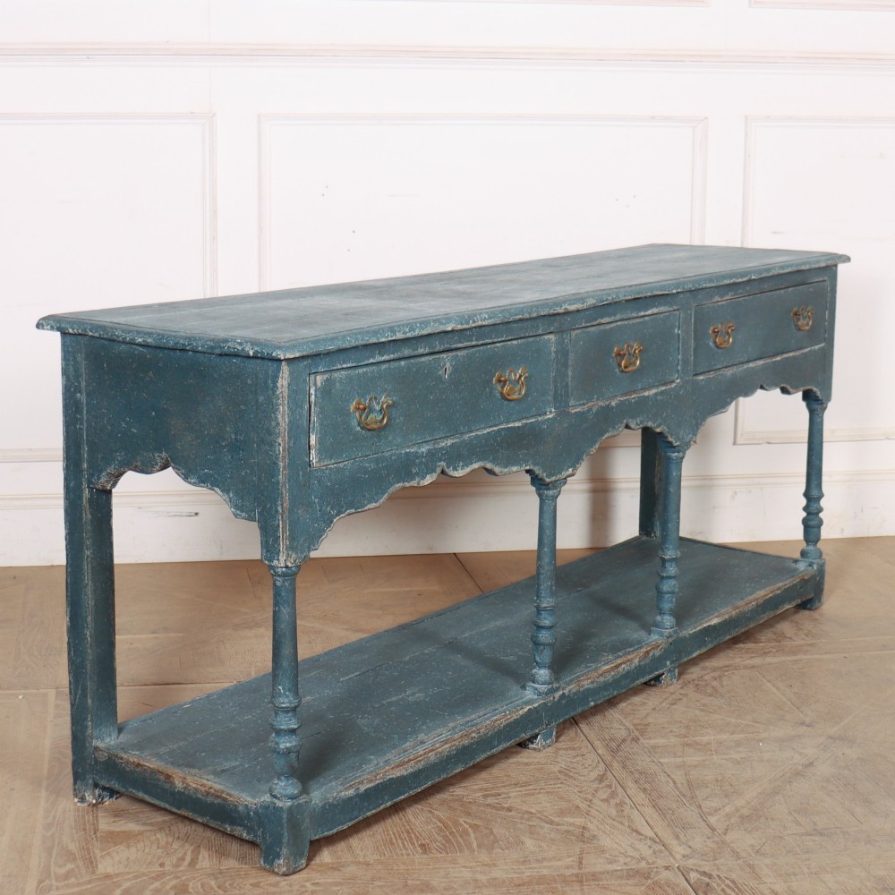 A long blue-painted wooden dresser base featuring three drawers with brass drop handles, a scalloped lower apron, turned front legs, and an open lower potboard shelf. View IX