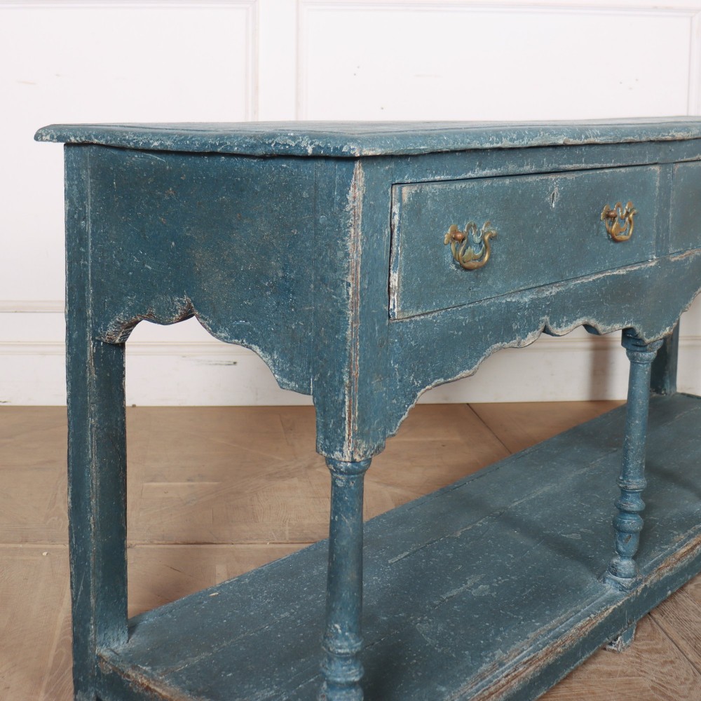 A long blue-painted wooden dresser base featuring three drawers with brass drop handles, a scalloped lower apron, turned front legs, and an open lower potboard shelf. View X