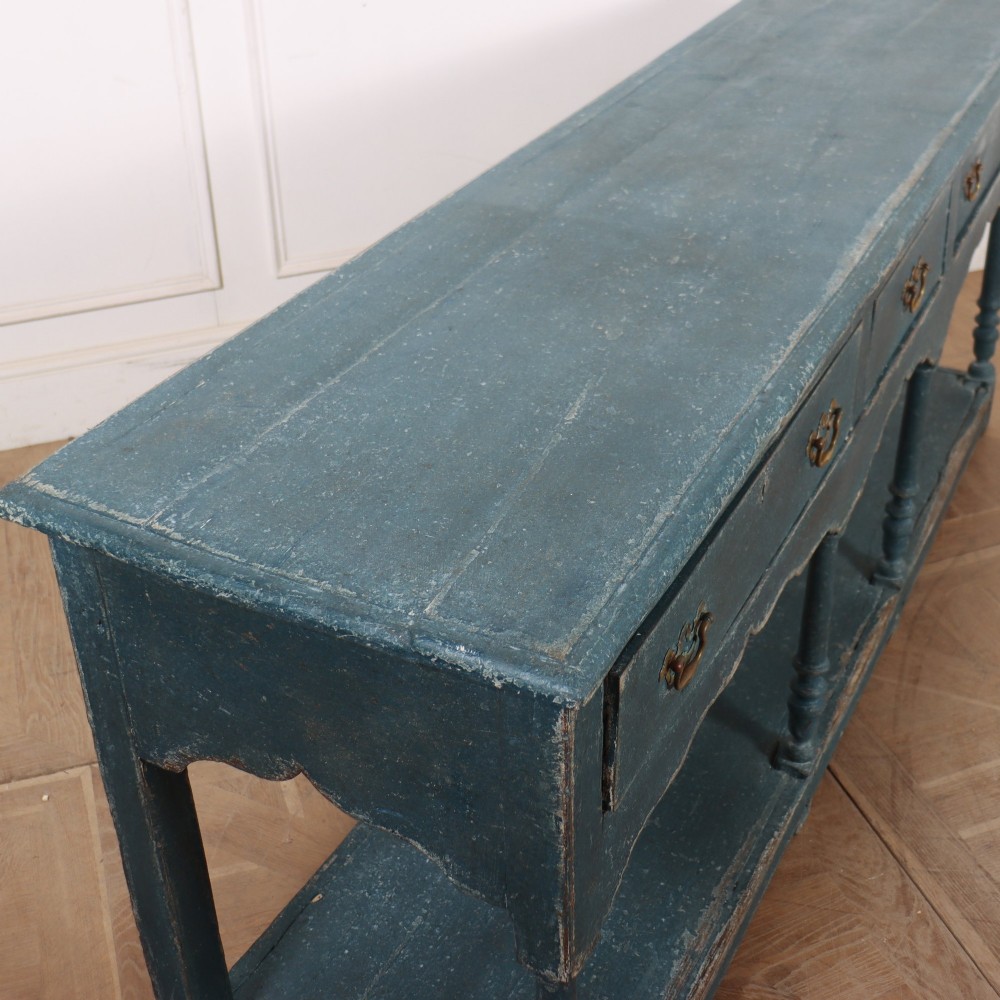A long blue-painted wooden dresser base featuring three drawers with brass drop handles, a scalloped lower apron, turned front legs, and an open lower potboard shelf. View XII