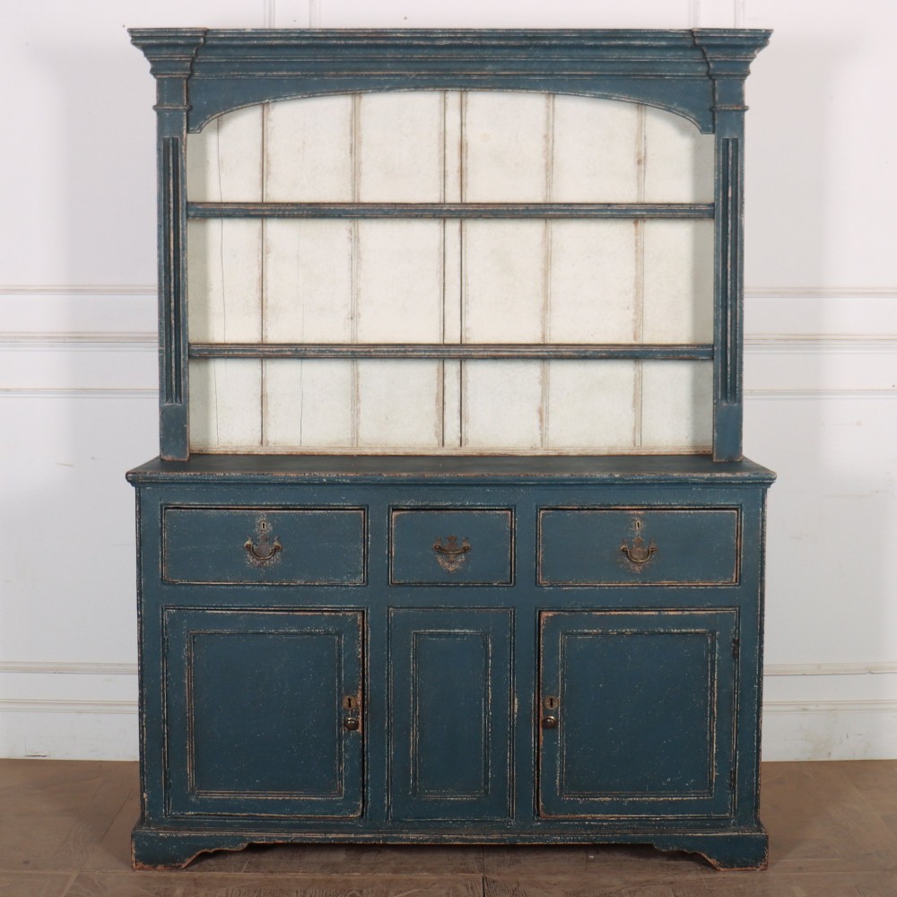 A large dark blue painted antique West Country dresser featuring an open plate rack with off-white backboards above three drawers and three panelled cupboard doors.