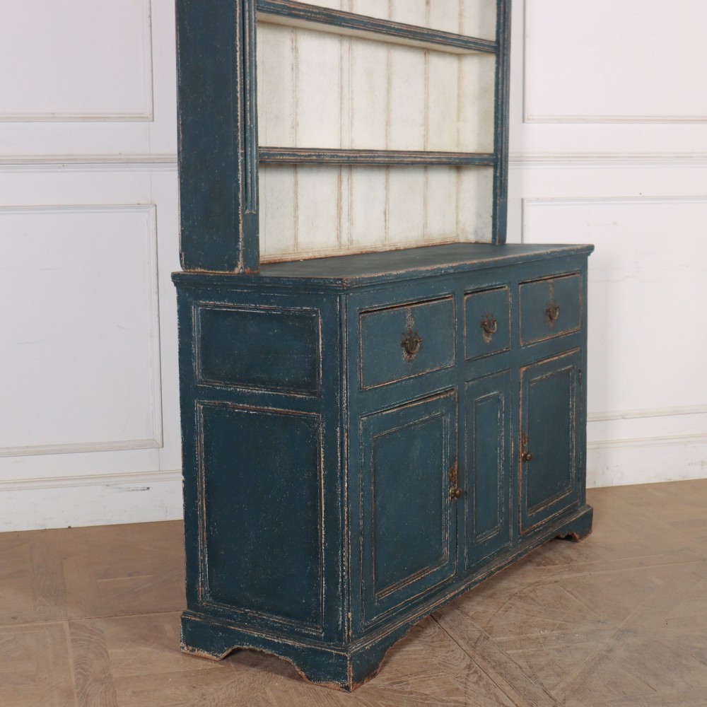 A large dark blue painted antique West Country dresser featuring an open plate rack with off-white backboards above three drawers and three panelled cupboard doors. View XI