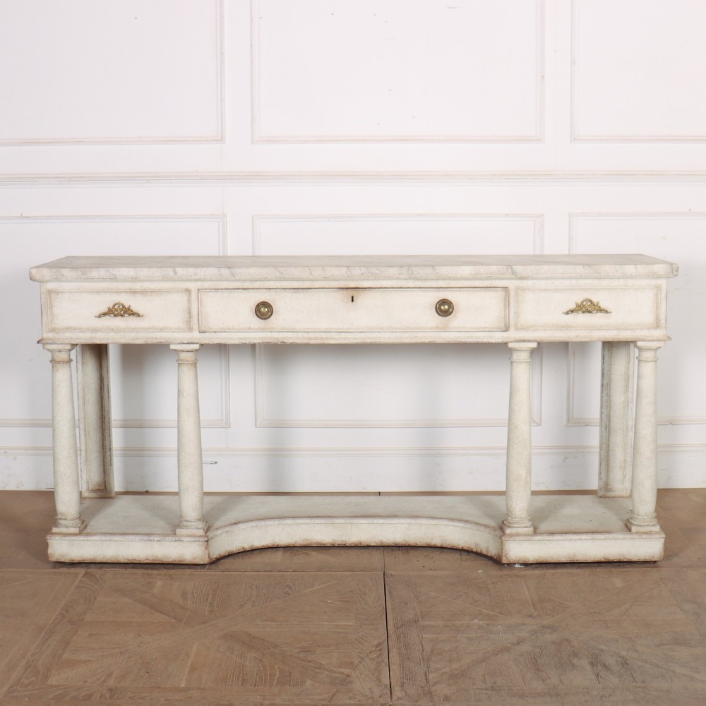 An antique off-white painted console table featuring a light marble top, three drawers with brass handles, four column supports, and a curved lower shelf.