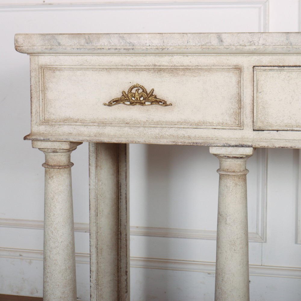 An antique off-white painted console table featuring a light marble top, three drawers with brass handles, four column supports, and a curved lower shelf. View II