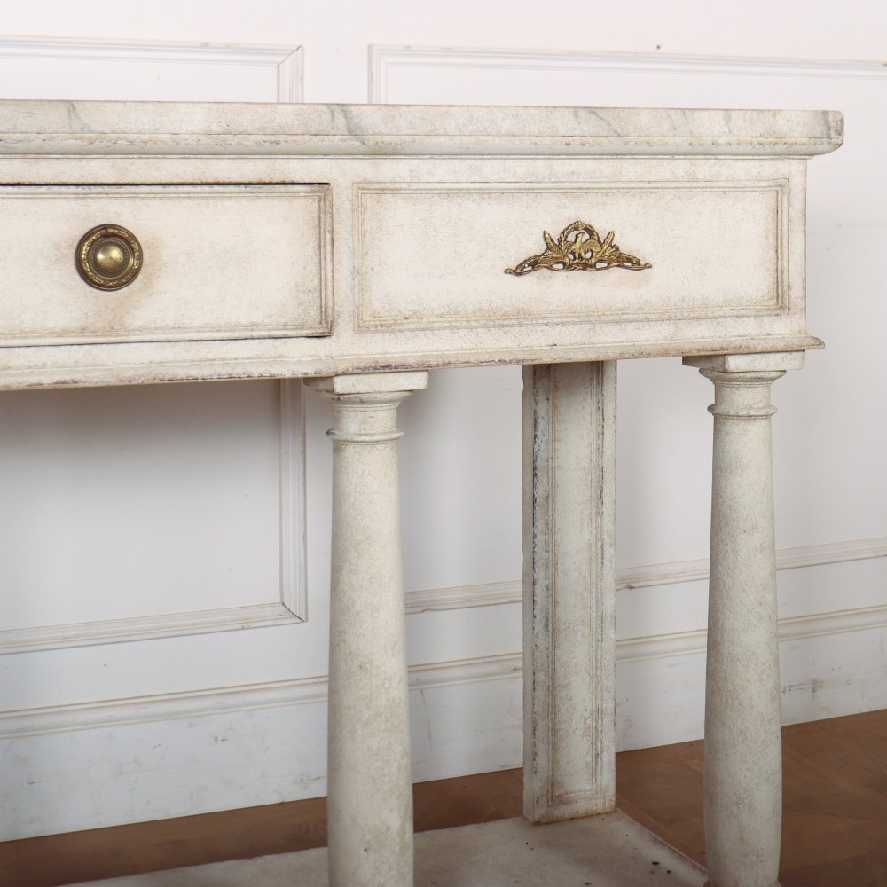 An antique off-white painted console table featuring a light marble top, three drawers with brass handles, four column supports, and a curved lower shelf. View IV